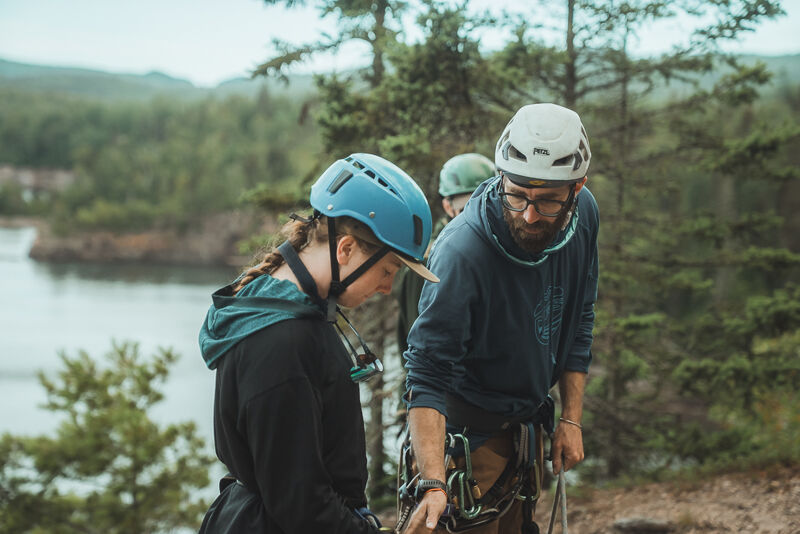 The image shows three people preparing for an outdoor activity, possibly rock climbing or rappelling. The main focus is on a person with a blue helmet, who is looking down and adjusting their gear. Another person, wearing a white helmet and glasses, is standing next to them, possibly assisting or instructing. A third person with a green helmet is standing behind them. They are surrounded by trees and a body of water is visible in the background, suggesting a natural environment.
